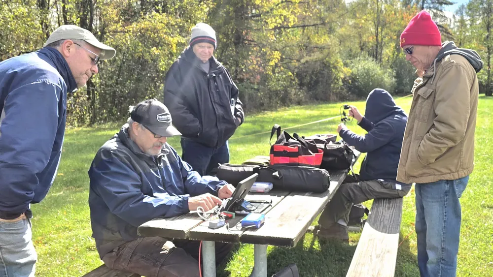 Members gather around a portable station at a picnic table in the park